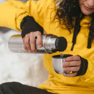 A woman pours a hot drink from a thermos into a metal cup while sitting in a snowy environment.