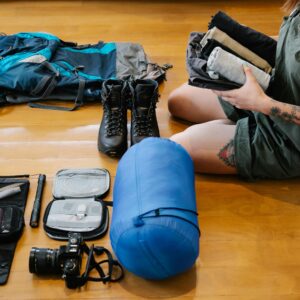 Woman organizing camping gear and clothes on wooden floor indoors, preparing for an adventure.