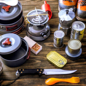 Camping cookware set with pots, pans, and utensils beside a campfire.